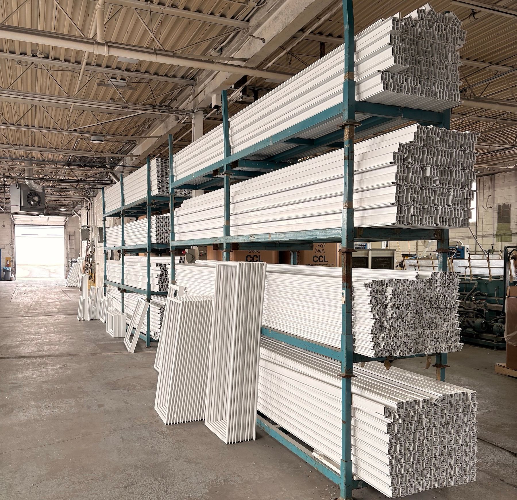 Industrial warehouse with metal shelving units holding stacks of long, white structural window or door frame components.