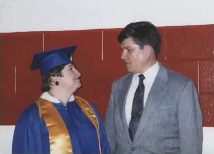 Graduation photo of two people in caps and gowns talking against a red wall