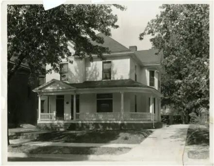 Black-and-white photo of a two-story house with a front porch and driveway, shaded by large trees