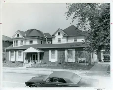 Black-and-white photo of a large house with a covered porch and a car parked in front