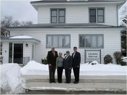 Four people standing in front of a white house in the snow, beside a sign reading “Lakeside Mortuary.”