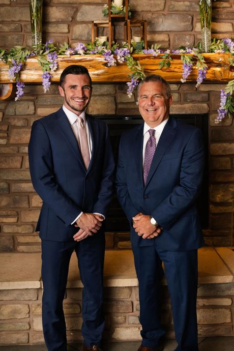 Two men in navy suits standing in front of a brick fireplace with flowers on the mantel