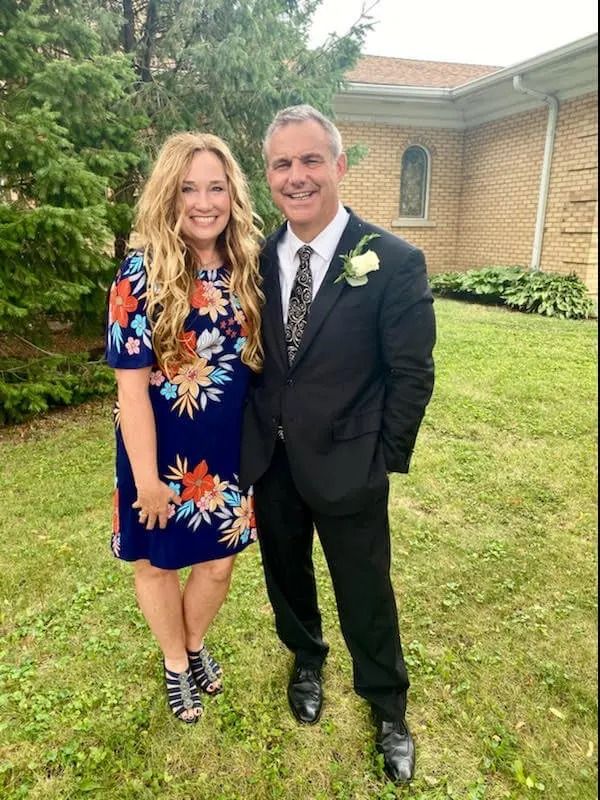 Two people smiling outdoors at a formal event, standing on grass before a brick building.