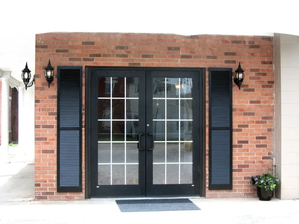 Brick storefront with black double glass doors, shutters, wall lamps, and a small potted plant.