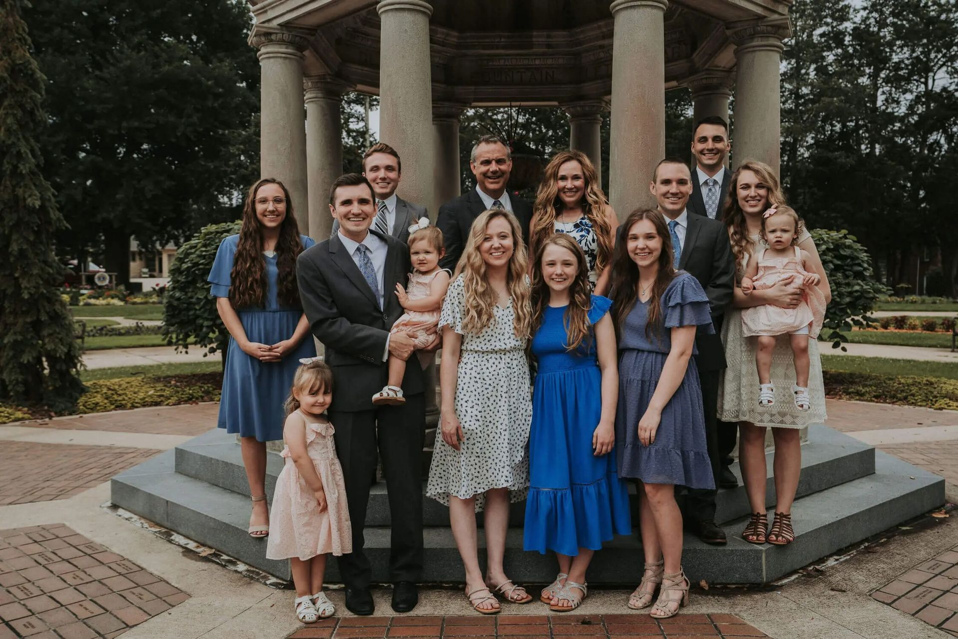 Large group posing together on steps in front of a gazebo, with several women in blue dresses and a man holding a baby.