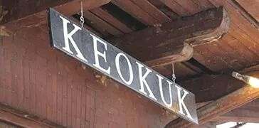 Black hanging sign with white letters reading “KEOKUK” under a wooden roof