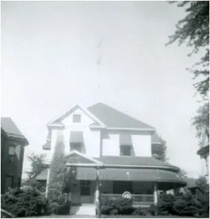 Black-and-white photo of a two-story house with a front porch and surrounding trees