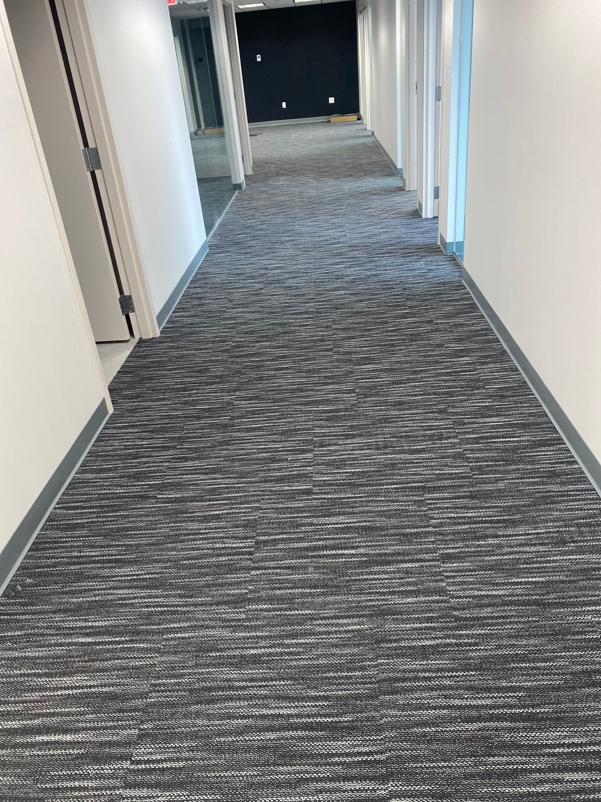 Empty office hallway with gray patterned carpet and white walls, lit by overhead lights.