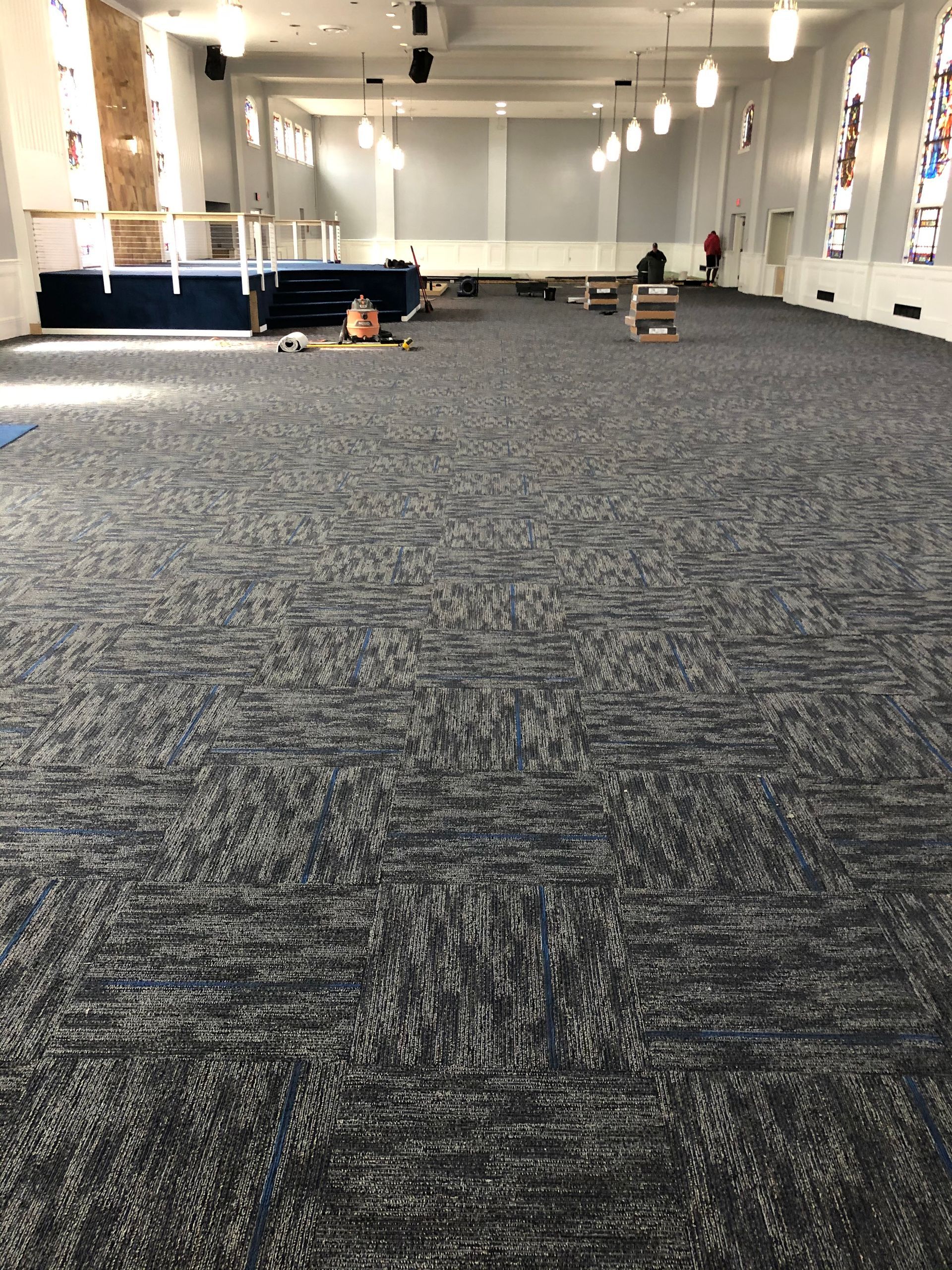 Empty carpeted indoor hall with white walls, tables along the sides, and a few scattered items in the distance
