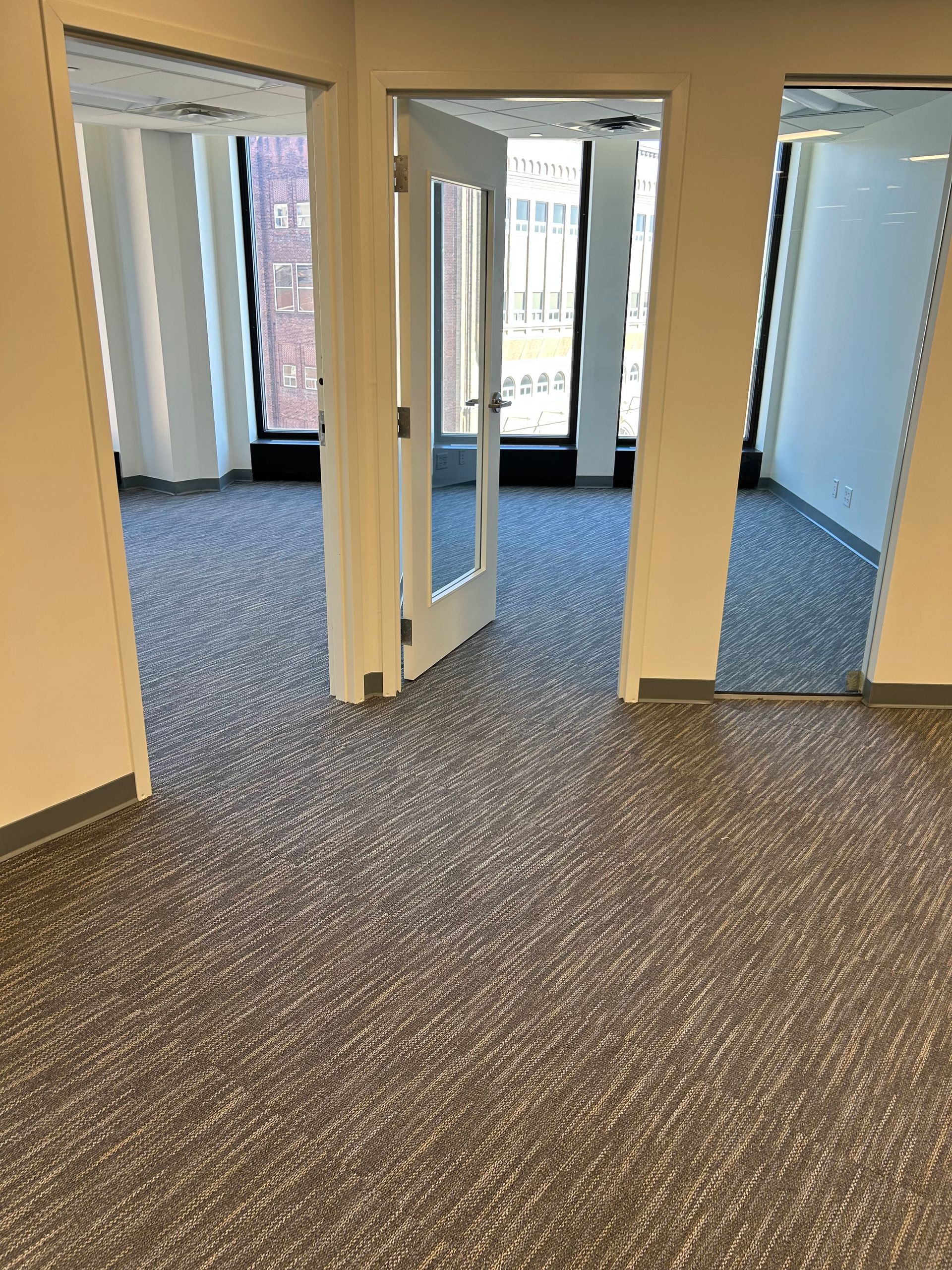 Empty office hallway with carpeted floors, beige walls, and open doorways leading to rooms with windows.