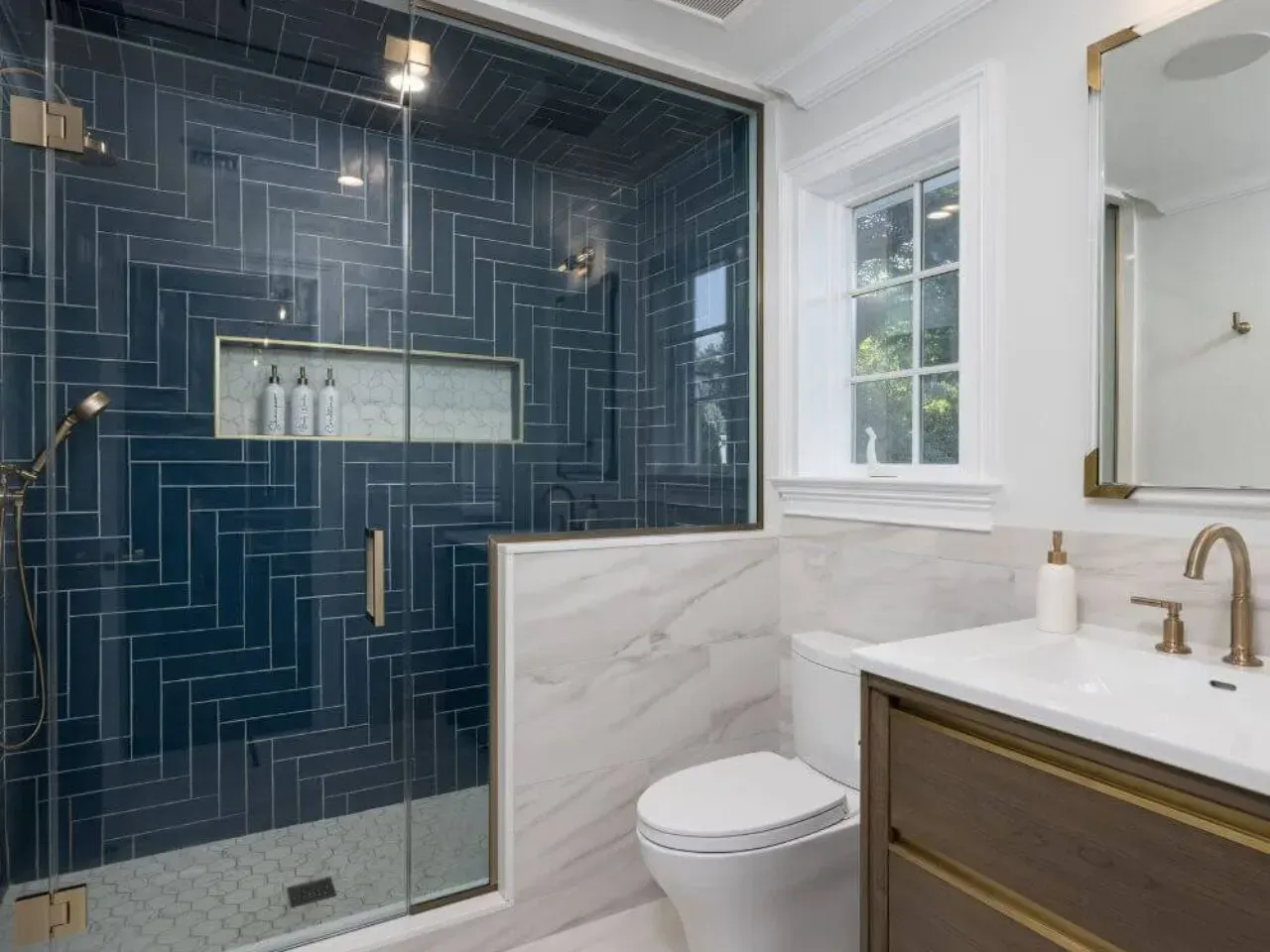Blue tile shower with glass door in modern bathroom. Toilet, sink, and window visible.