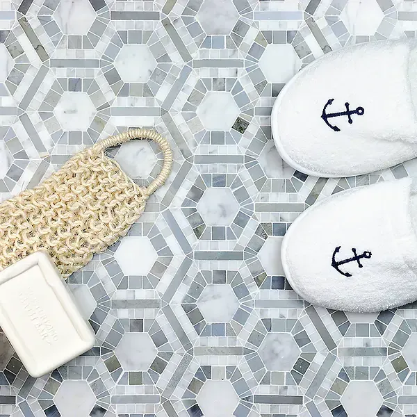 White slippers with anchor embroidery next to soap and a body scrub on a geometric tile floor.