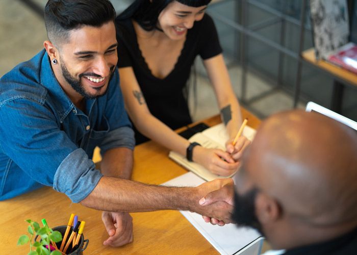 Lender and clients smiling and shaking hands at a table with papers and pencils