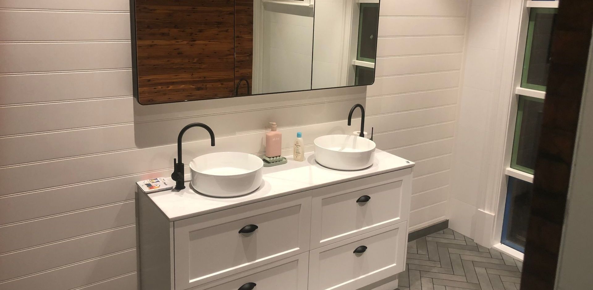 Bathroom with white sink cabinets, vessel sinks, and wood-paneled mirror above.