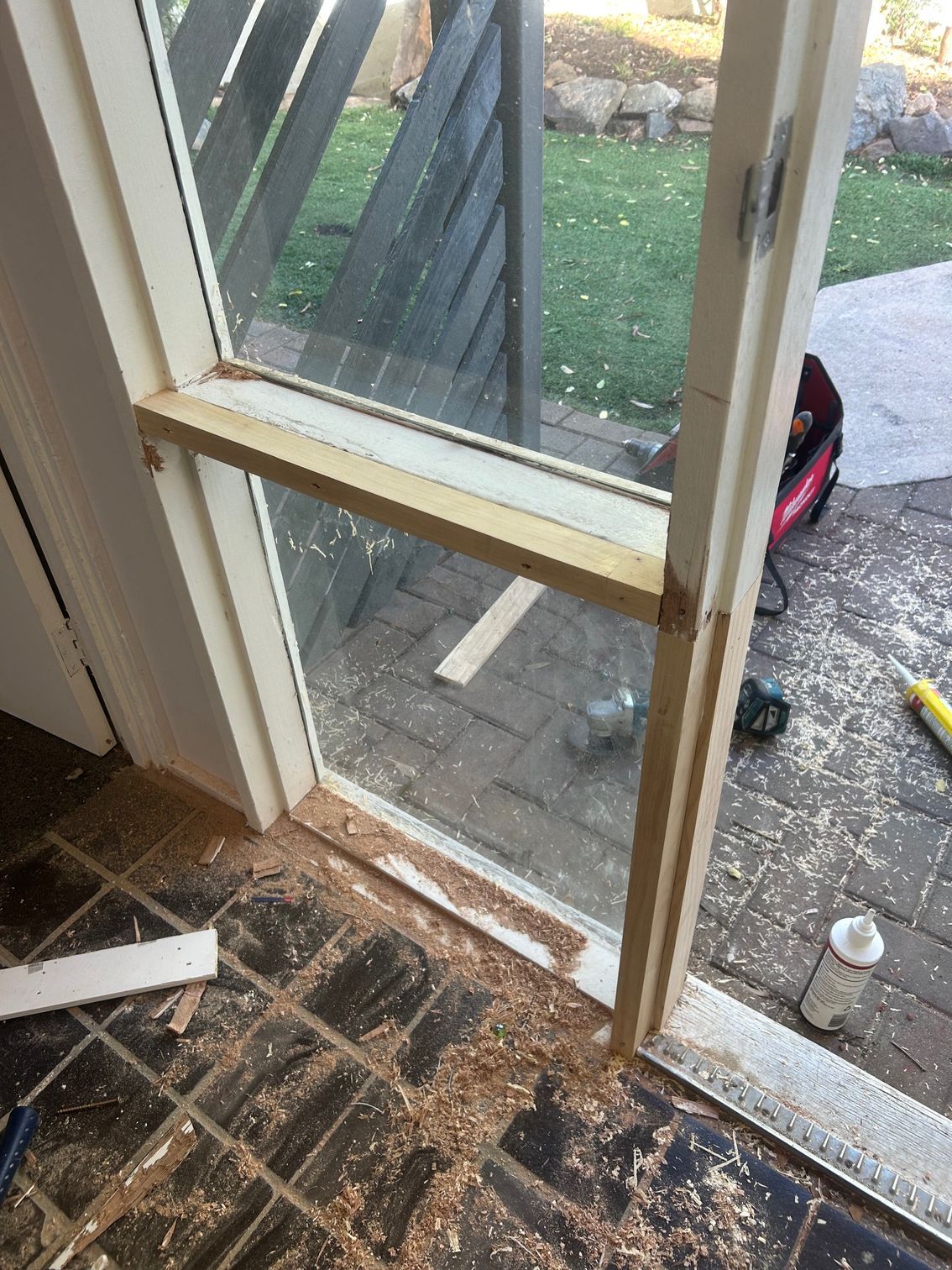 Window frame being repaired; wood shavings on tiled floor; sunlight outdoors.