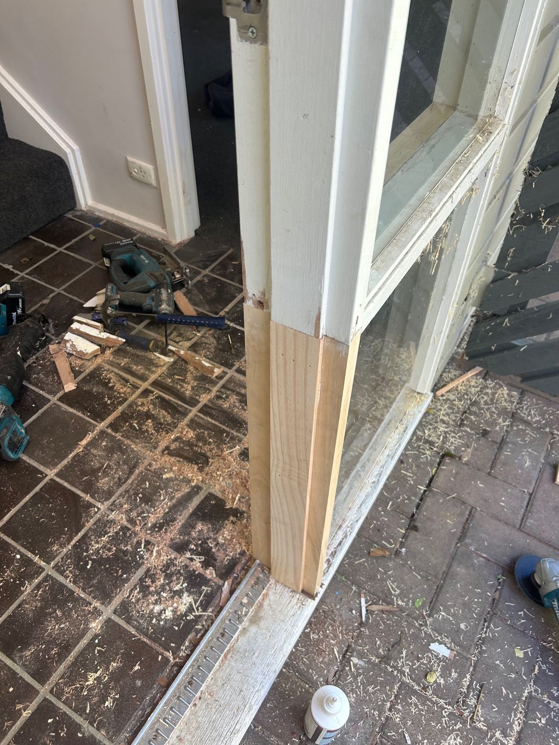 Door frame being repaired; wooden trim partially installed. Tools and wood shavings on tiled floor.