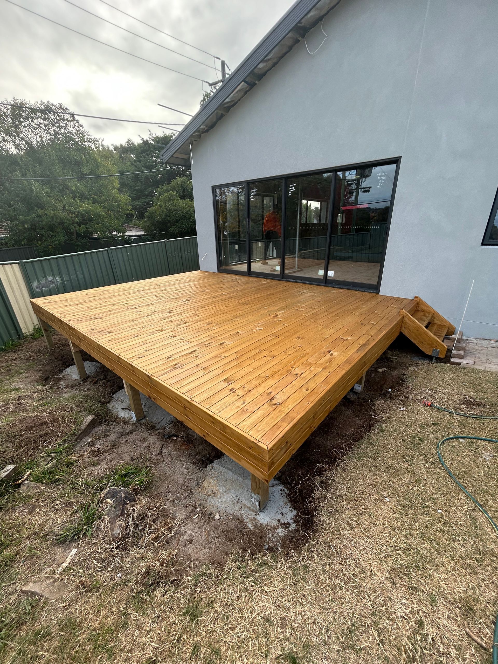 Wooden deck outside a house, with steps, overlooking a yard. The deck is supported by concrete blocks.