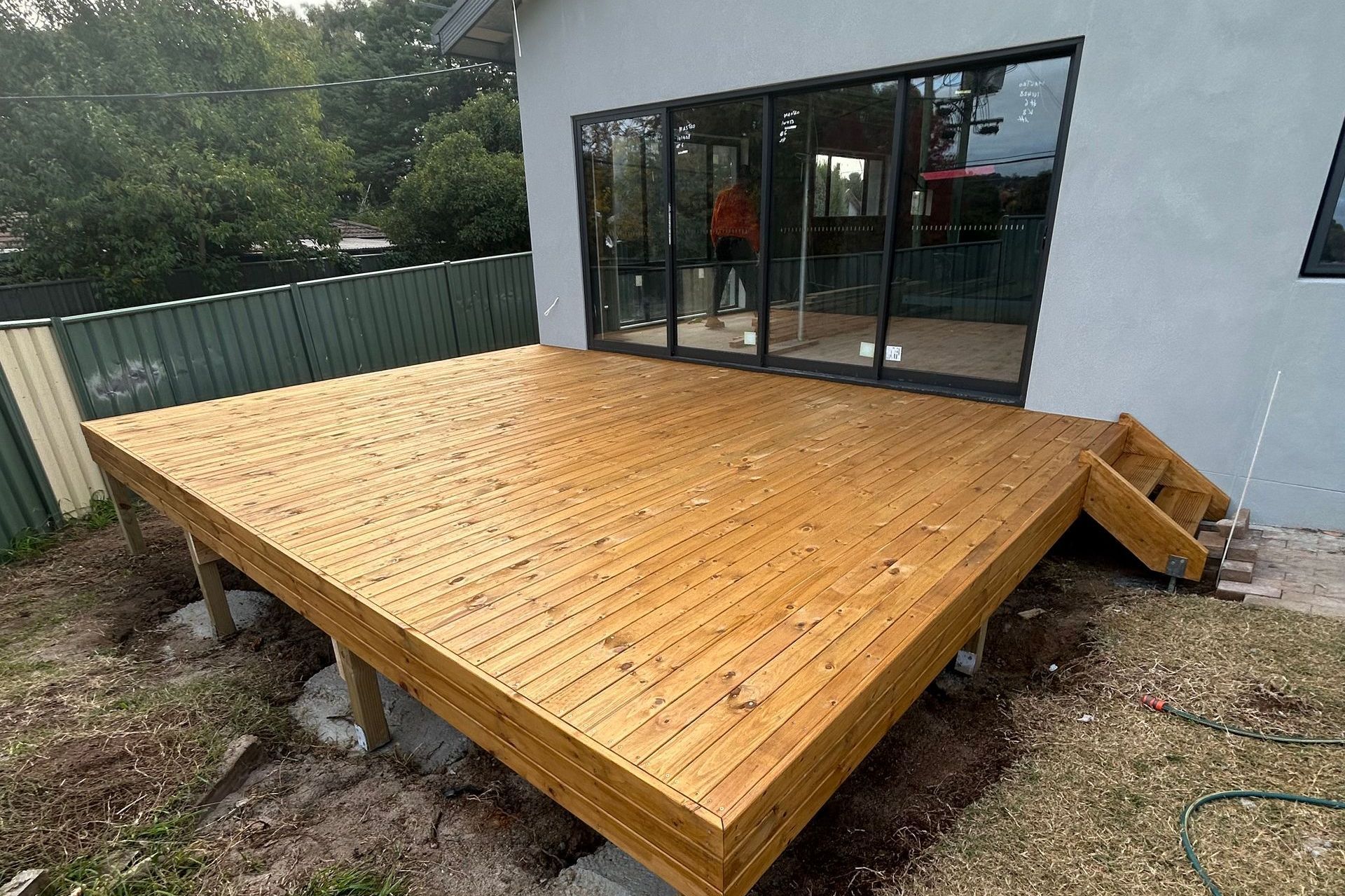 Wooden deck outside a house with glass sliding doors. Stairs lead down to ground.
