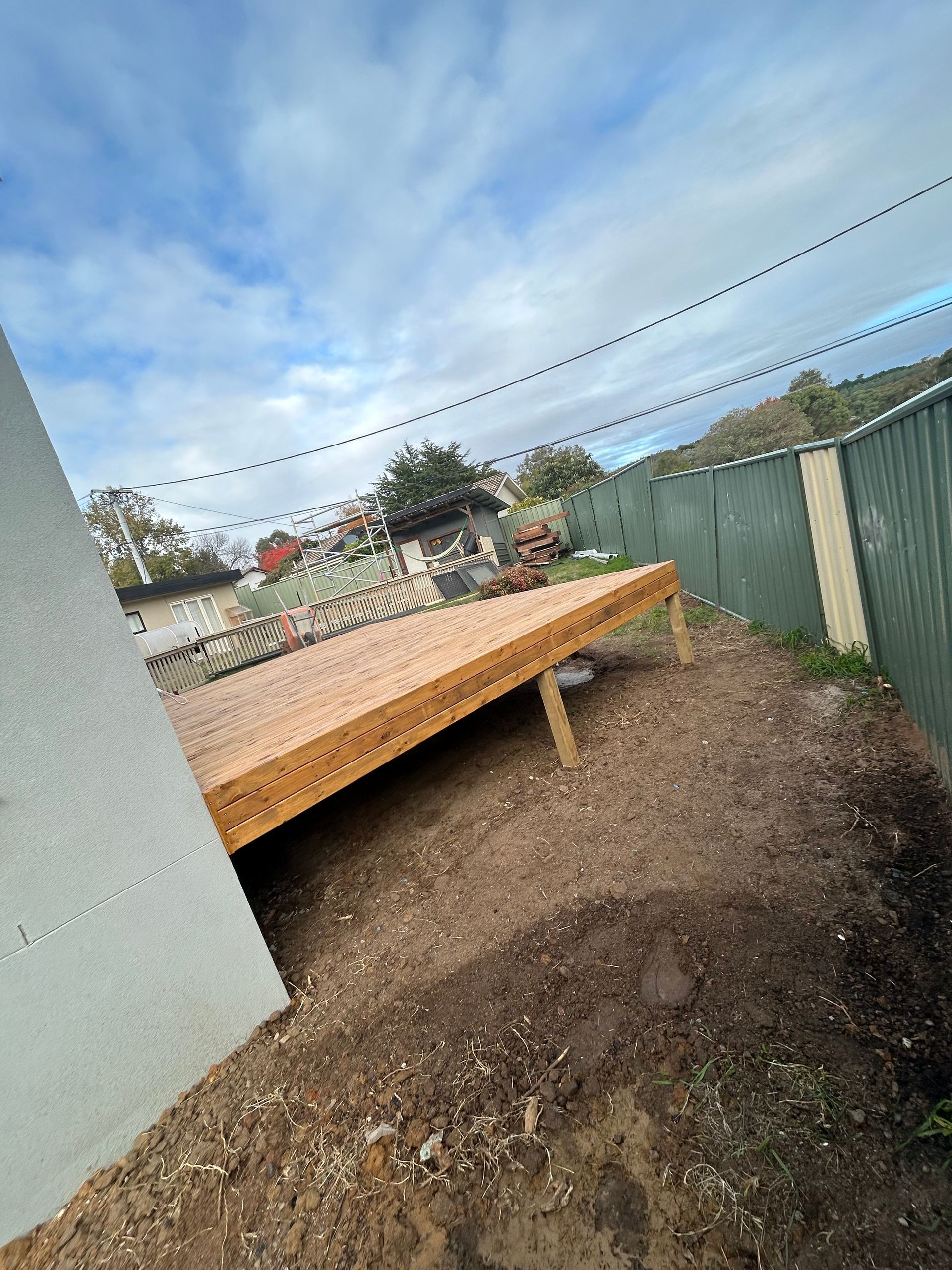 Wooden deck under construction next to a fence and a building, cloudy sky above.