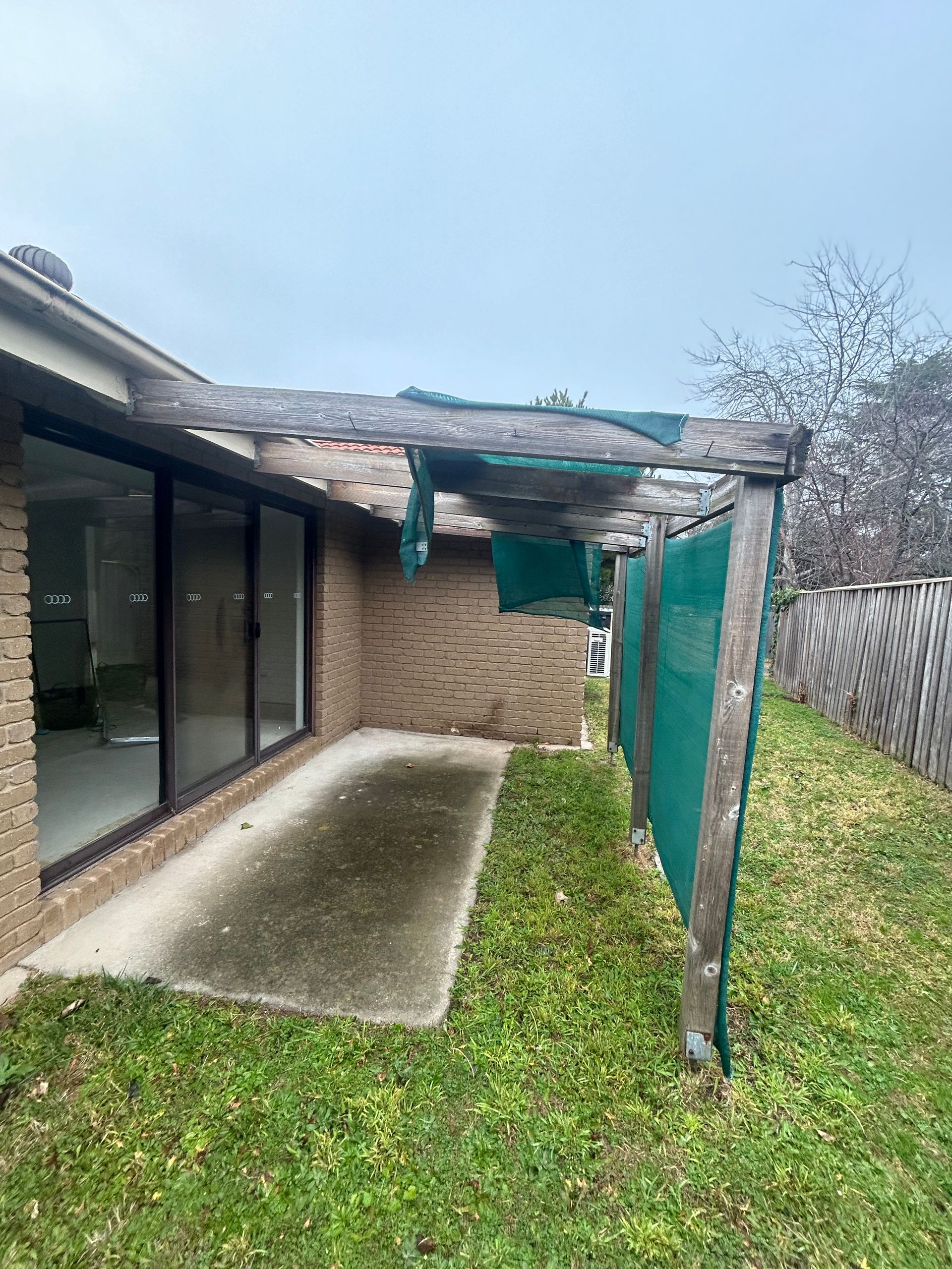 Patio area with weathered brick wall, concrete floor, green shade cloth, and wooden support beams. Overcast day.