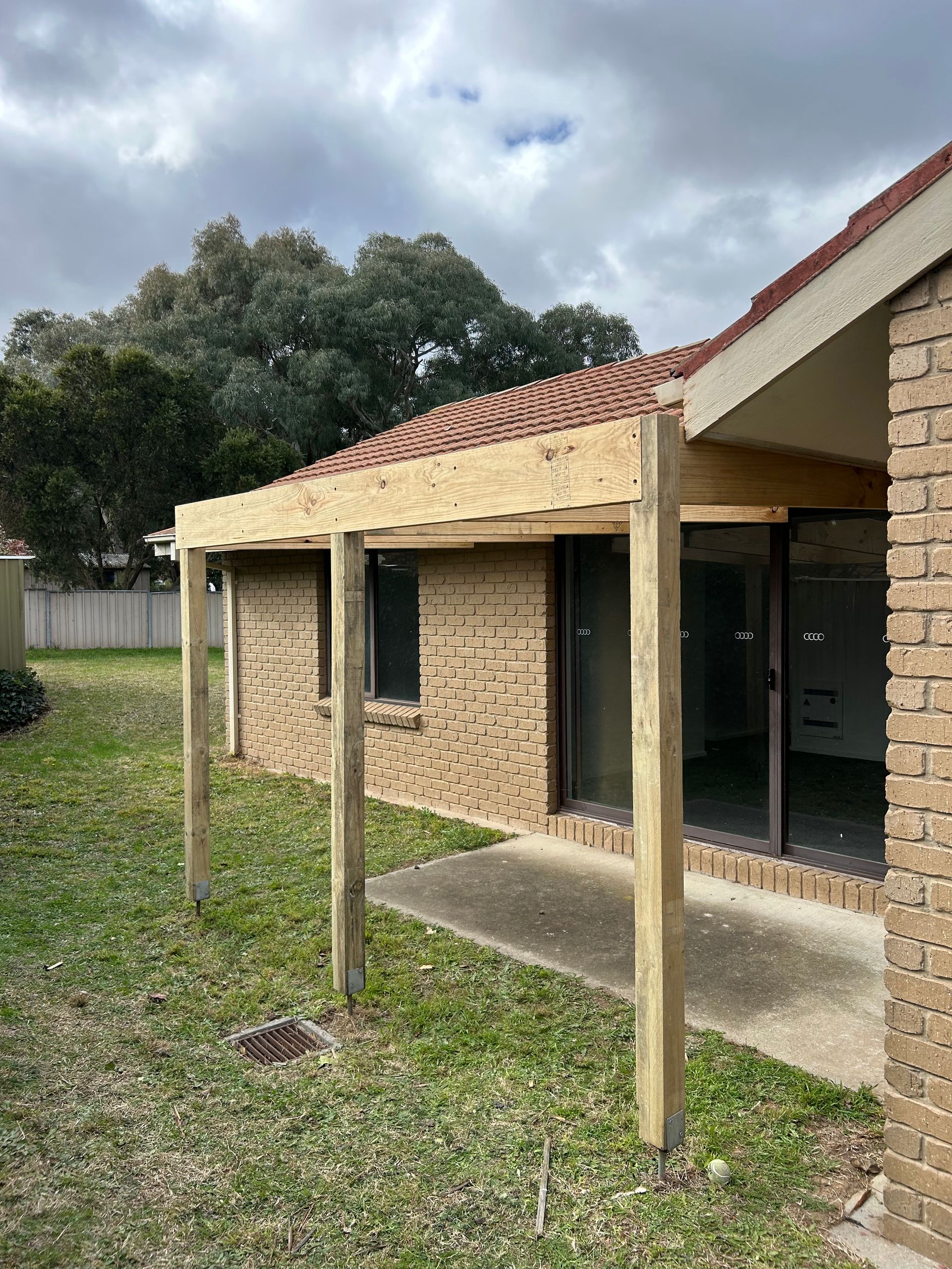 A wooden patio under construction attached to a brick house. Green lawn and overcast sky.