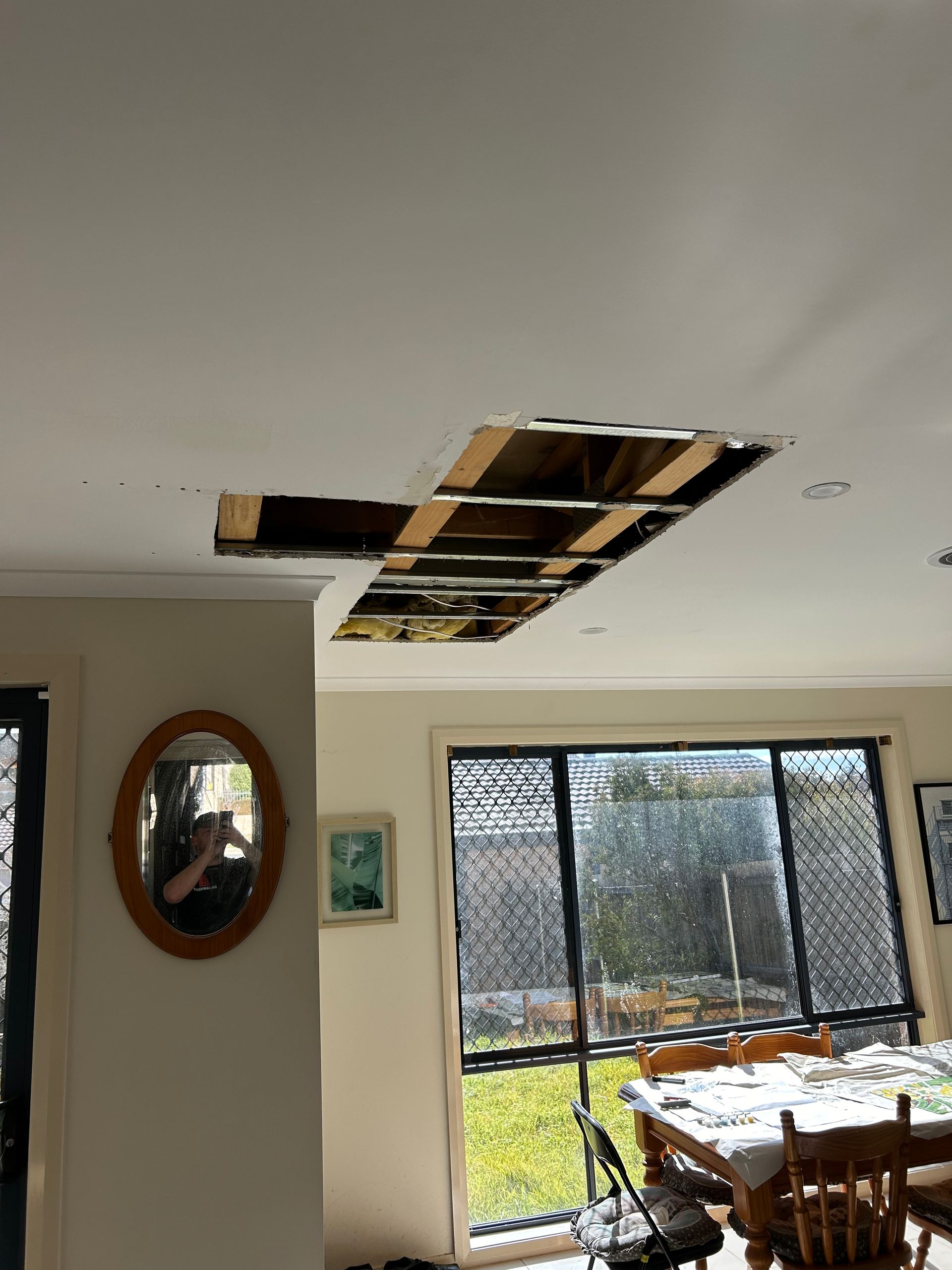 Ceiling damage with a large rectangular hole exposing wooden beams, overlooking a dining area with a window.
