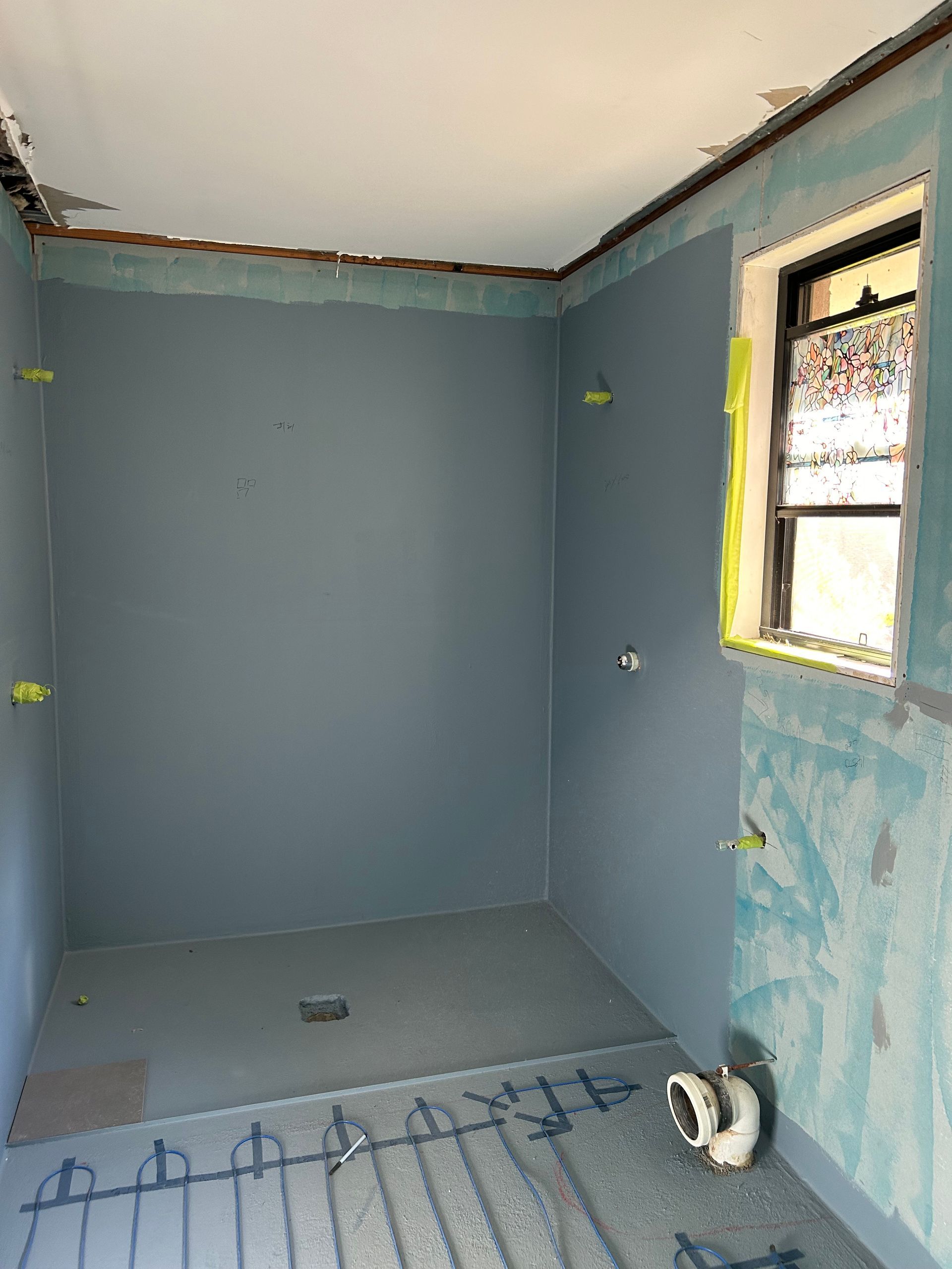 Bathroom under construction, blue waterproof walls, gray floor, window on right. Pipes visible.