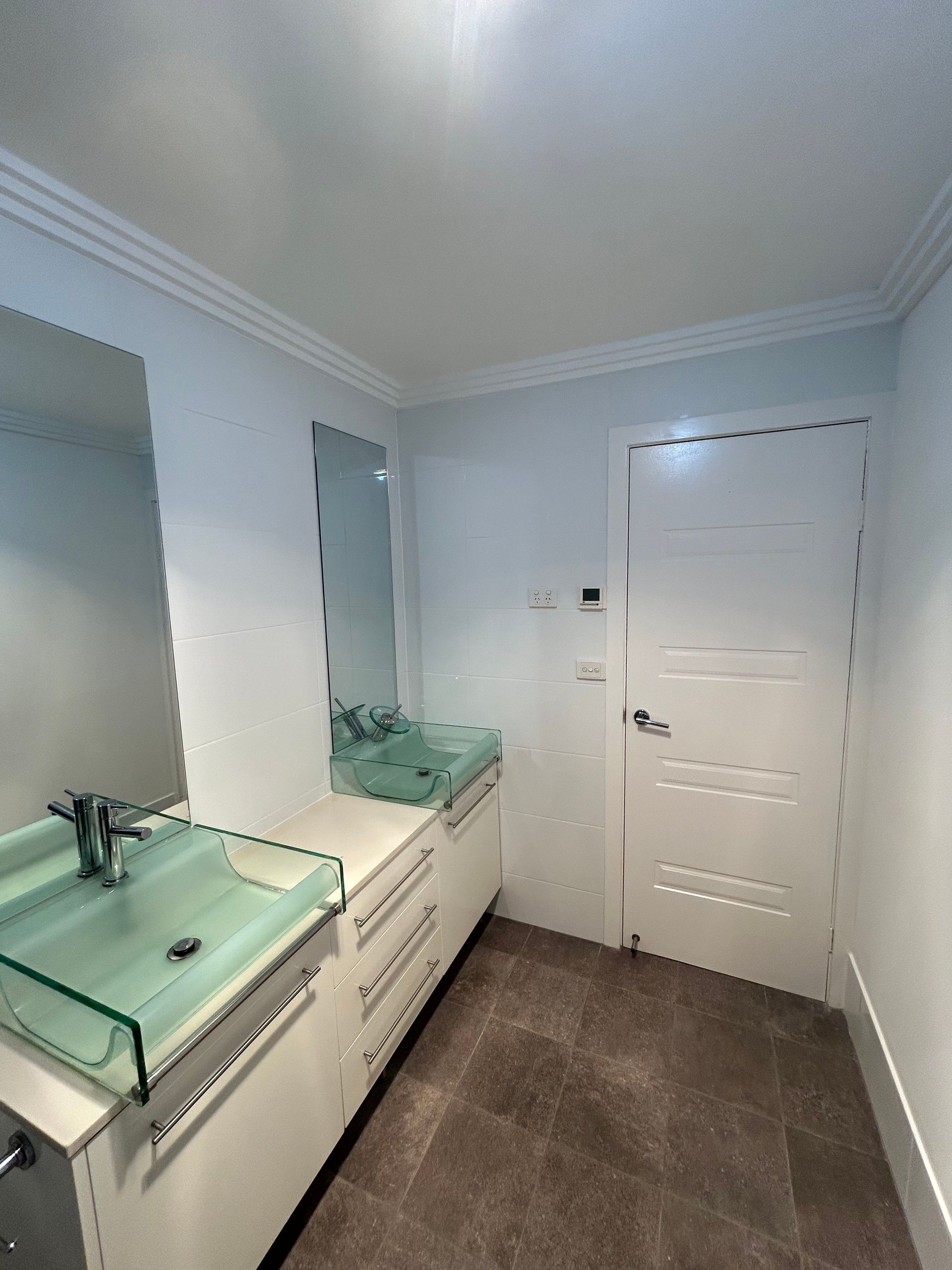 Bathroom with white walls, cabinets, and a glass sink. A door is on the right, and the floor is brown tile.