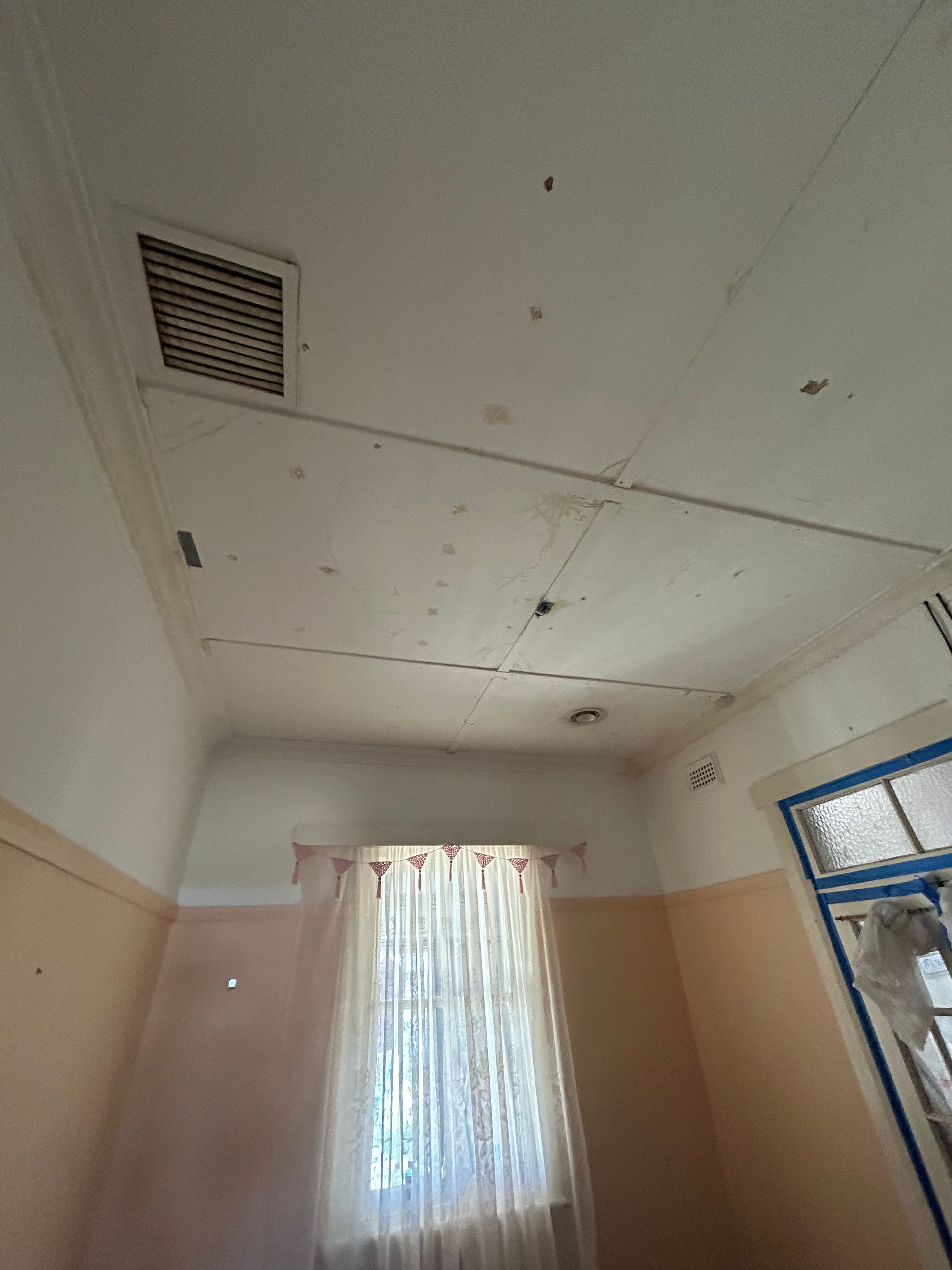 Interior room with damaged ceiling, air vent, window with sheer curtains, and light-colored walls.
