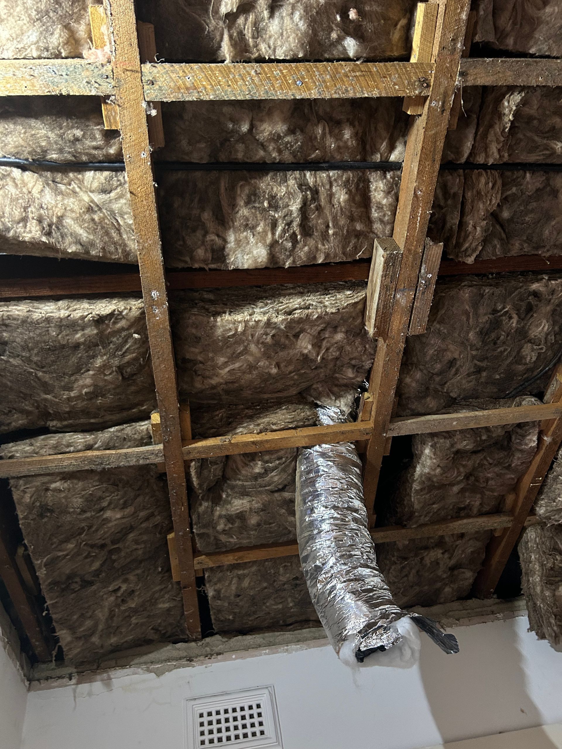 Attic interior showing wood beams, insulation, and a metal duct; small white vent in the wall.