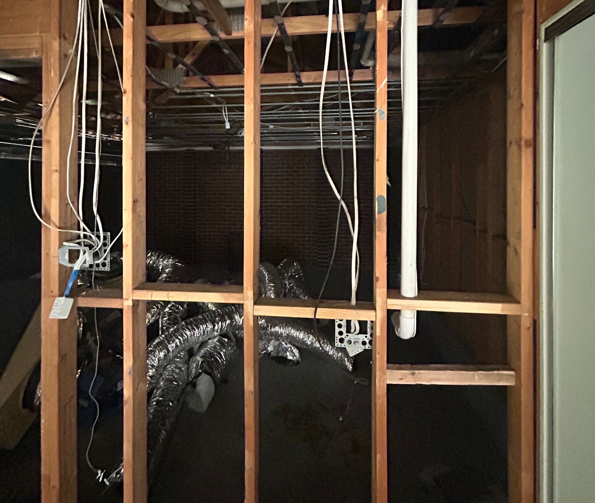 Wooden wall frame with exposed wiring, pipes, and ductwork. Brick wall visible in background.