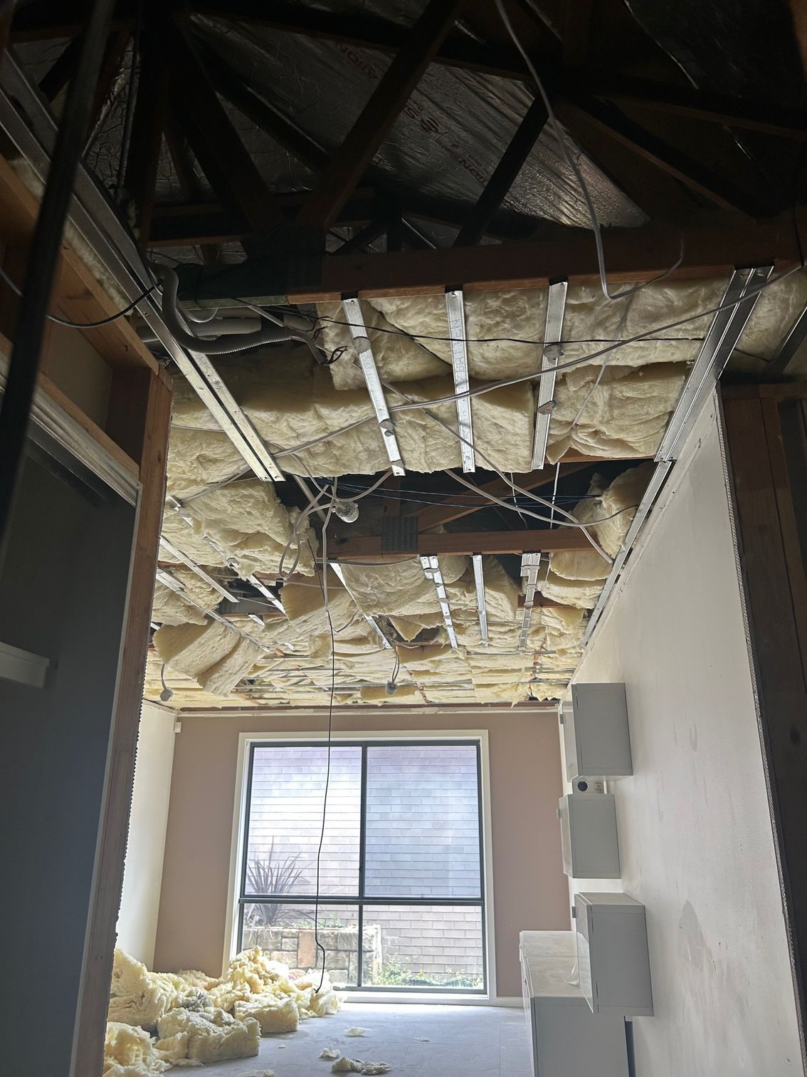 Interior view of a damaged ceiling with exposed beams, insulation, and a window in the background.
