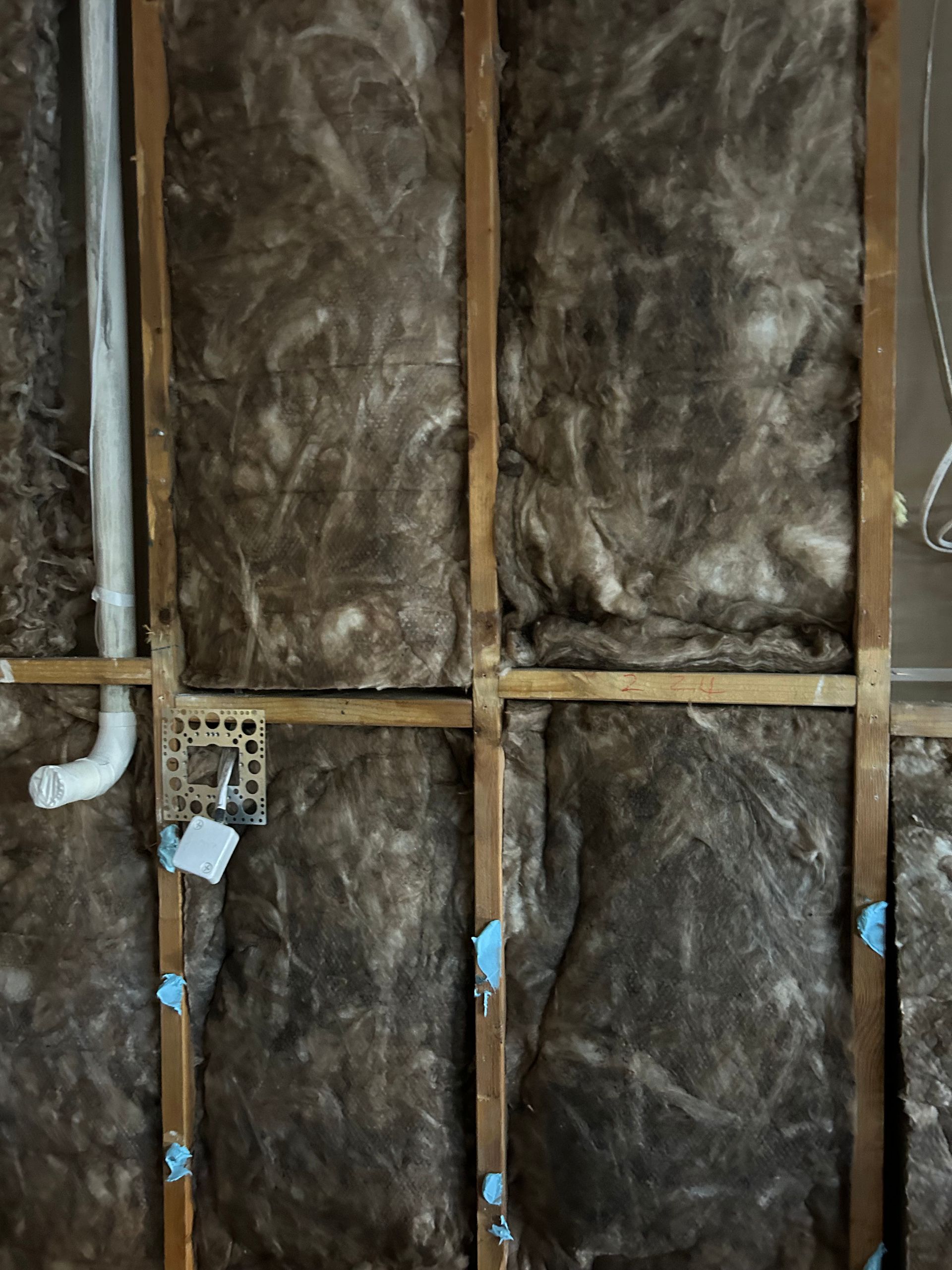 Wooden wall framing with brown insulation, electrical outlet, and white plumbing.