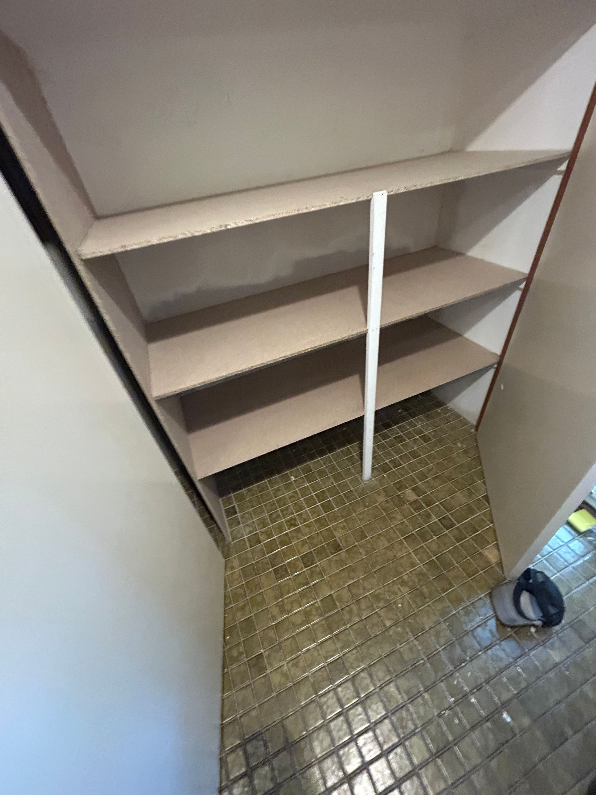 Empty closet with shelves, supported by a central white pole. Beige shelves, white and brown trim, tiled floor.
