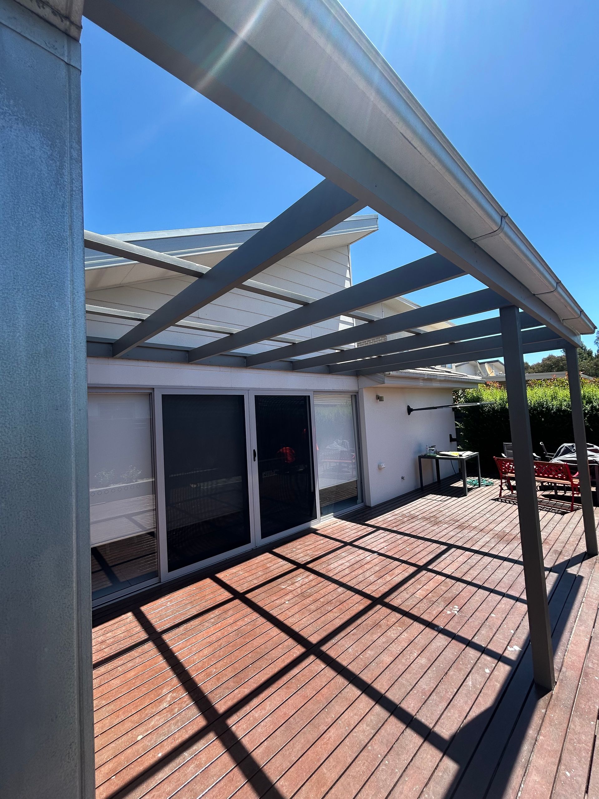 Deck with overhead beams casting shadows on the wooden planks, leading to glass doors of a white building on a sunny day.