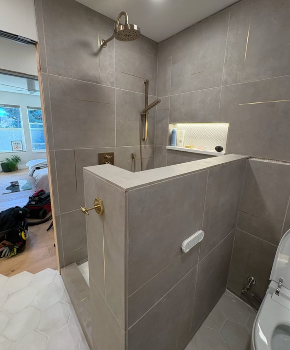 Modern bathroom with gray tiled shower and toilet, gold fixtures, and built-in shelf.