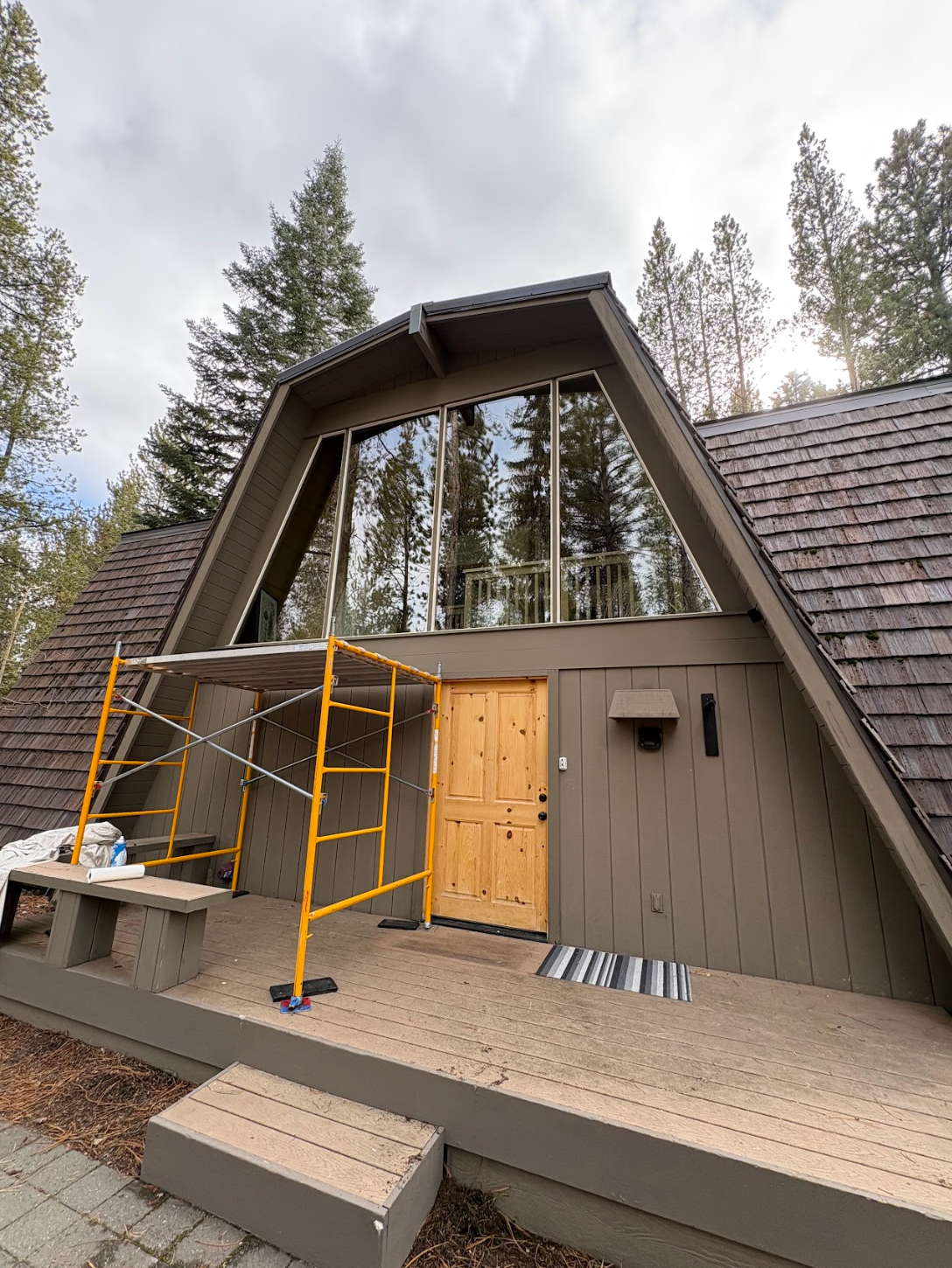 A small cabin under construction with yellow scaffolding, wood siding, and tall trees in the background.