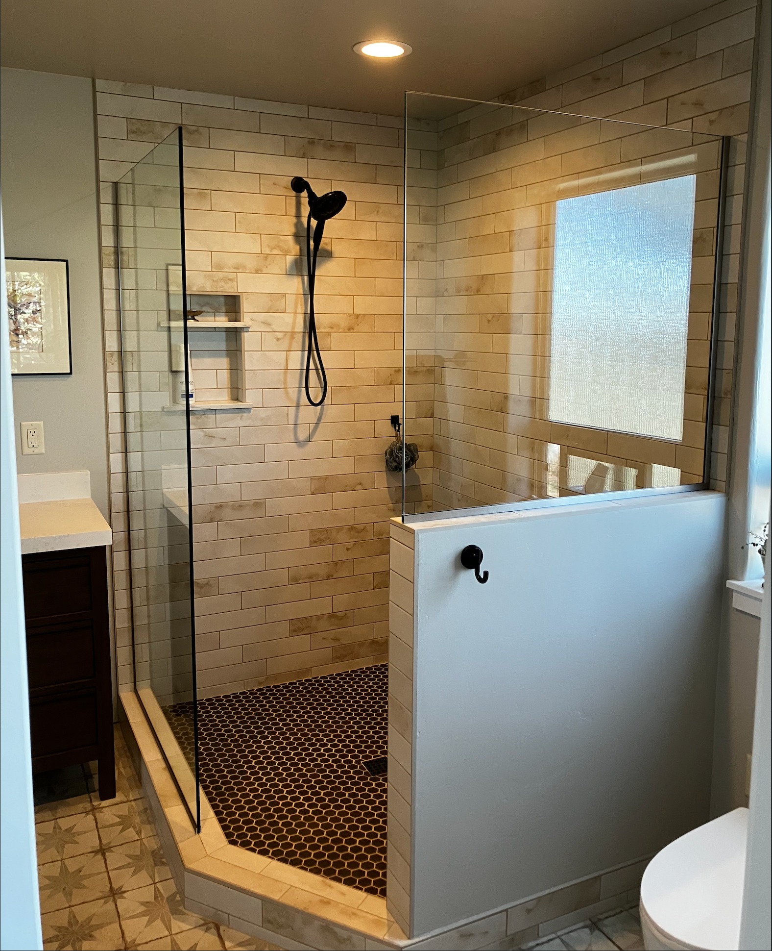 A modern walk-in shower with stone tile walls, a glass enclosure, patterned floor tiles, and a small built-in shelf.