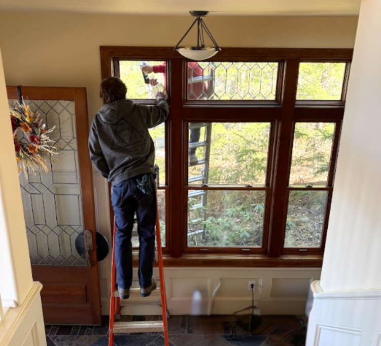 A person on a ladder cleans a large, multi-pane wooden window inside a house.