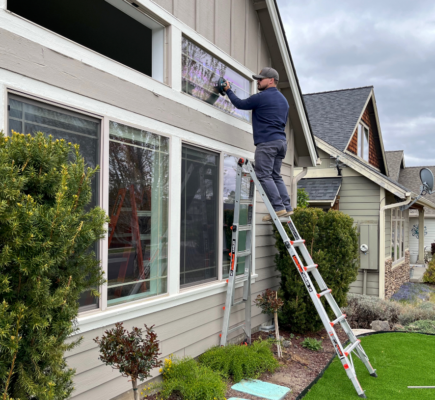 Man on a ladder cleaning a window of a light-colored house. Green grass and bushes surround the house.