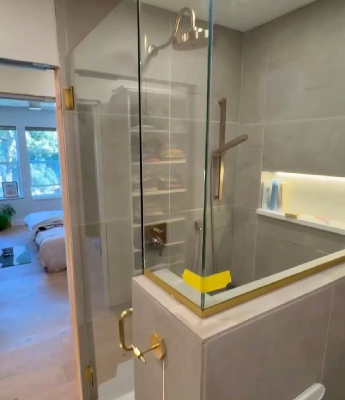 Modern shower with gold fixtures and glass doors; a bedroom is visible in the background.