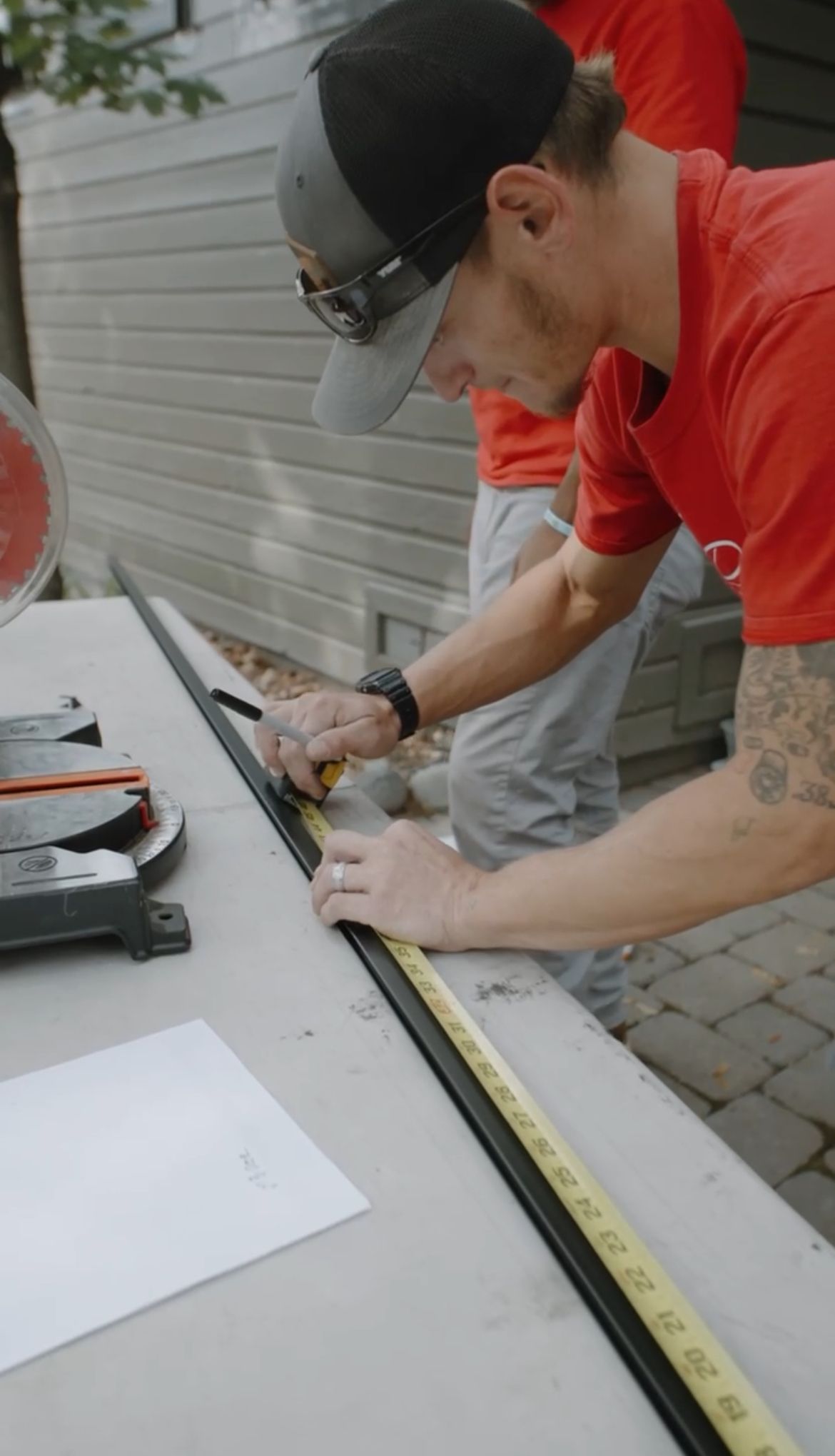 Man in red shirt uses a utility knife to measure and cut a long black piece on a table outdoors.