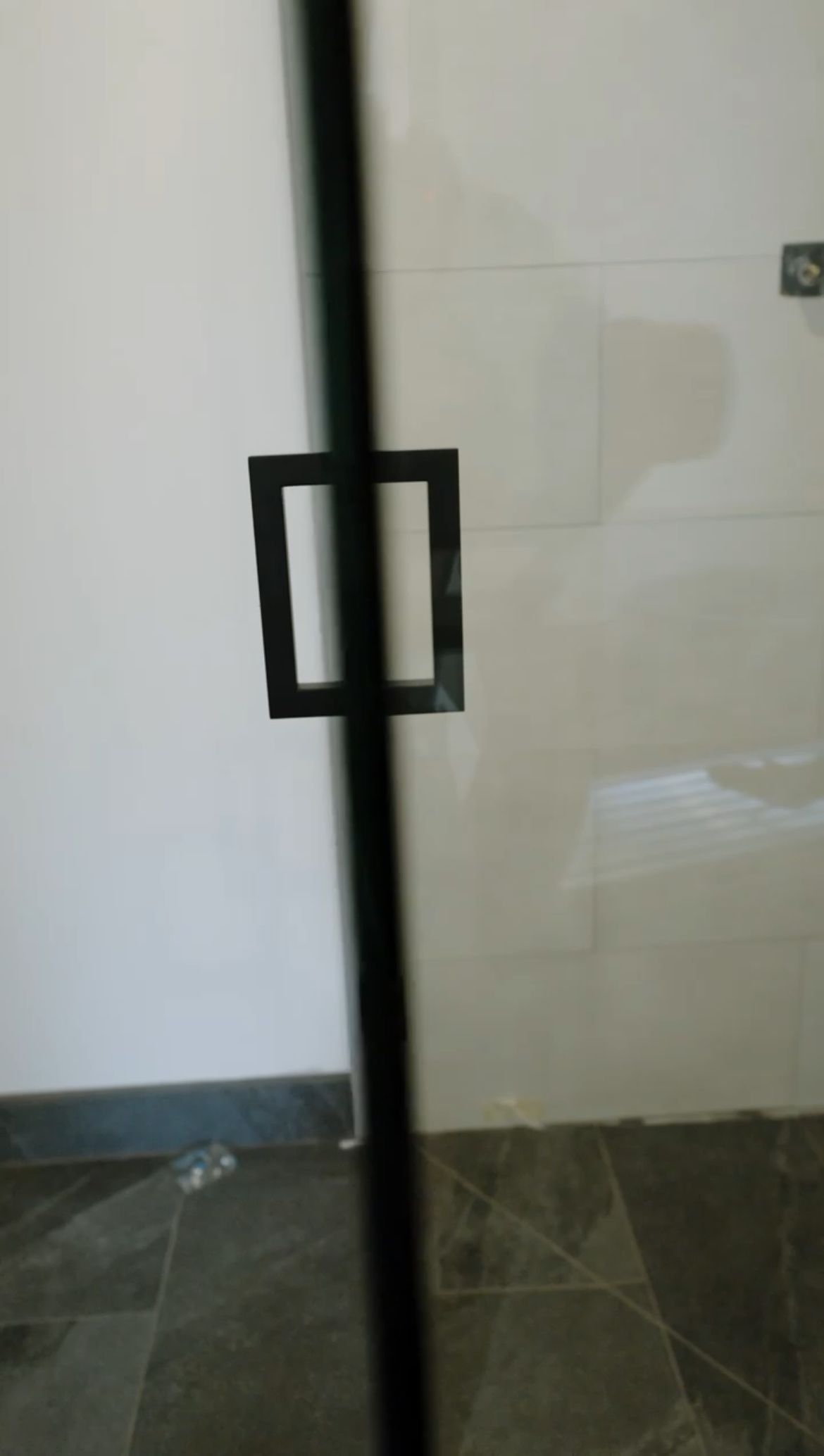 Black square handle on a glass shower door against a white wall and dark floor.