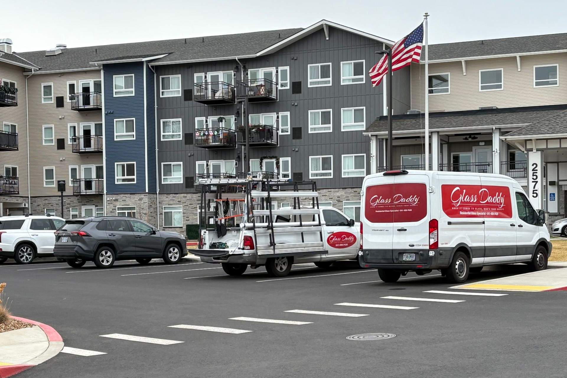 Vehicles parked in front of an apartment building, including a white van with red logos. American flag flying.