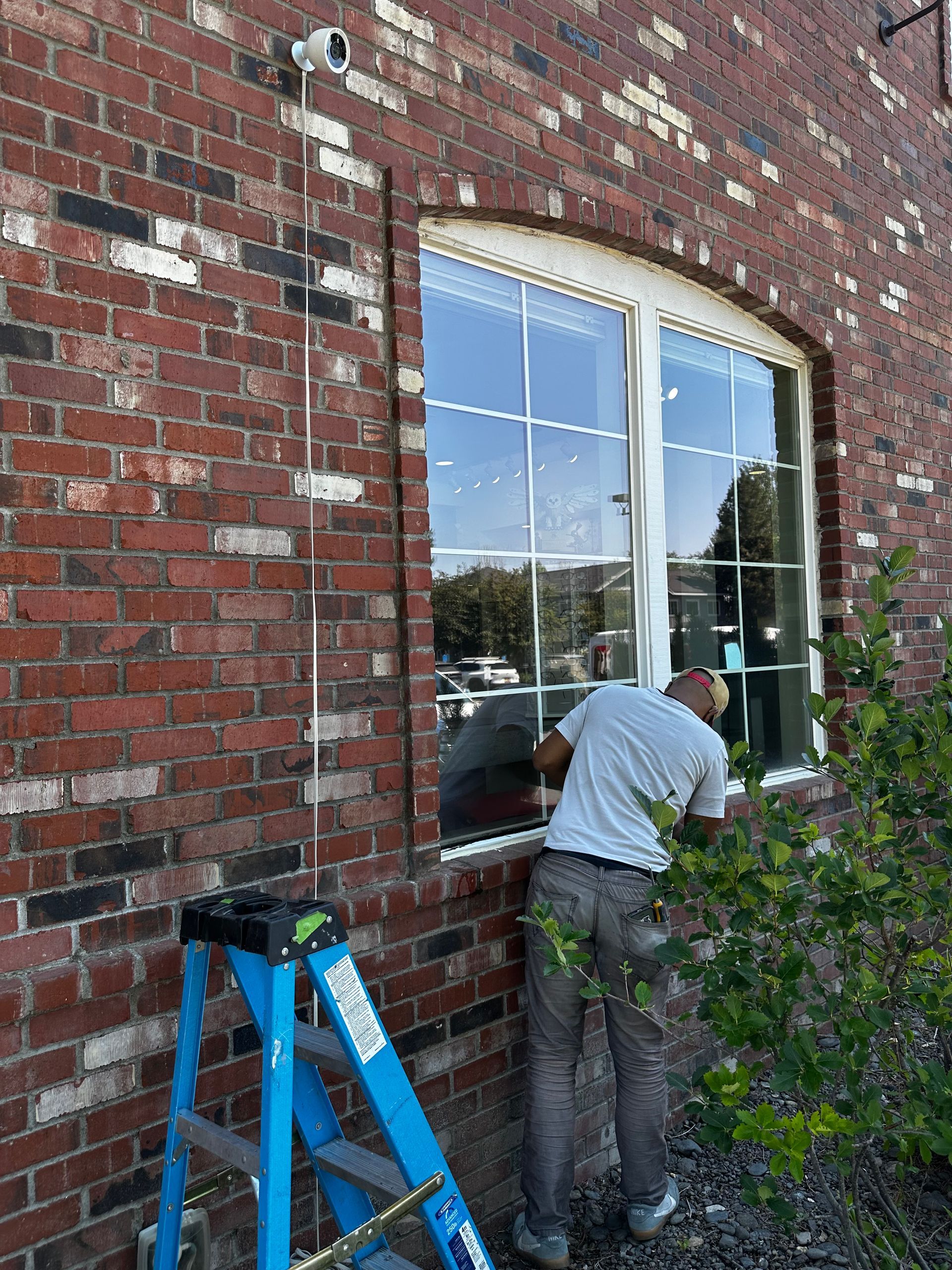 Person repairing a brick wall beside a large window, with a blue ladder nearby.