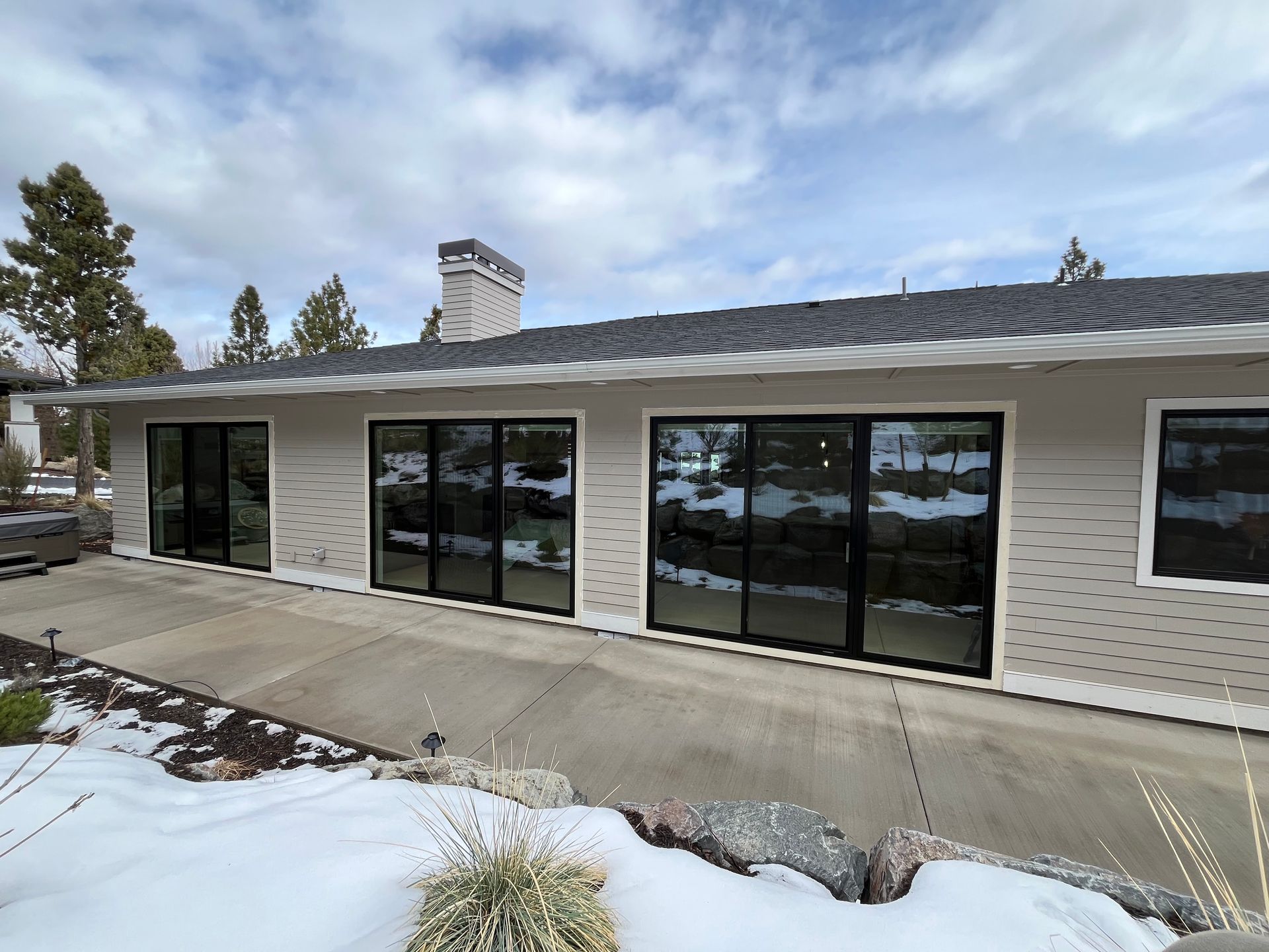 Beige house with black-framed windows, concrete patio, and snowy ground under a cloudy sky.
