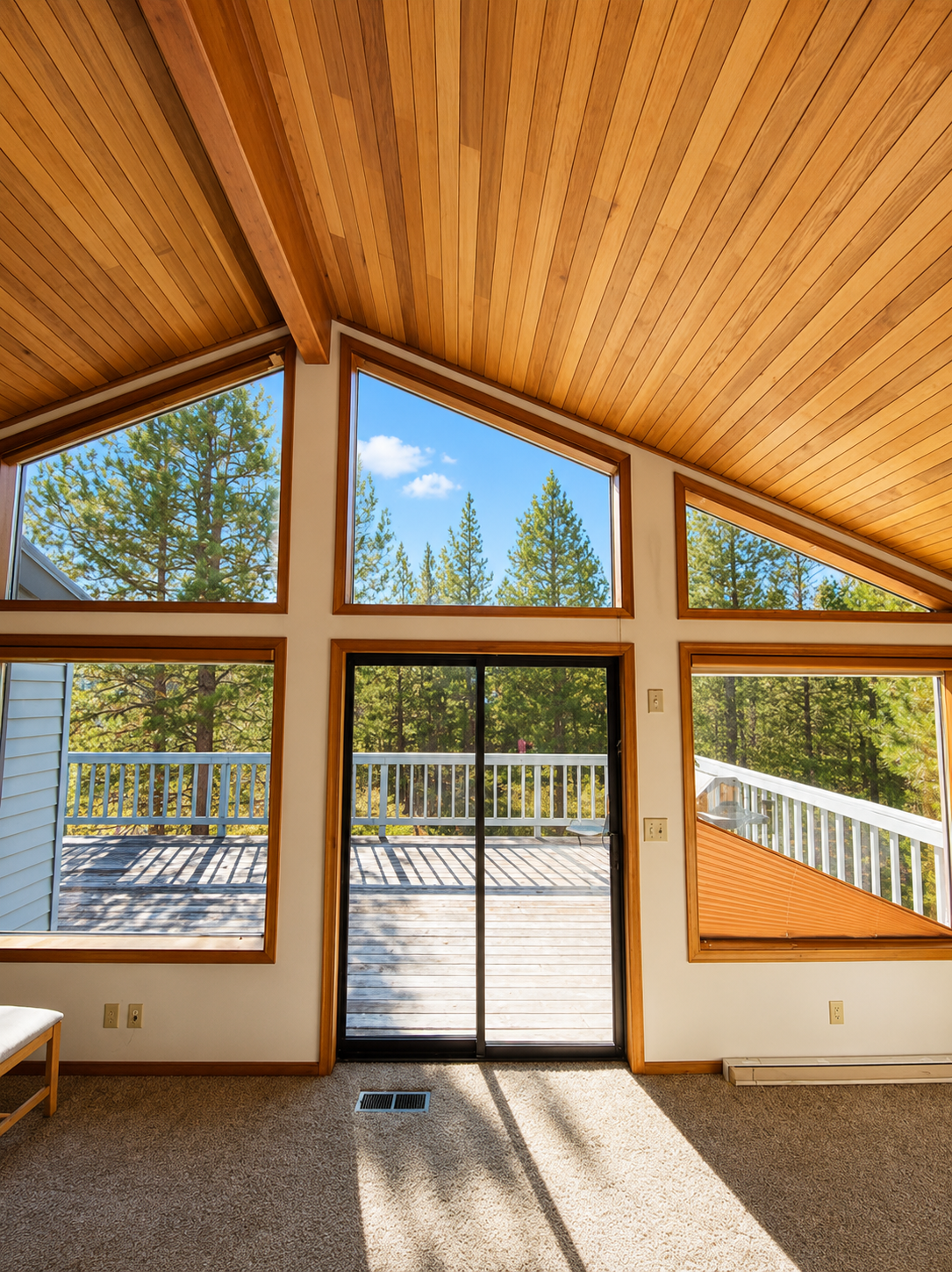 Vaulted wood-ceiling room with tall windows and glass doors opening to a sunlit deck and trees.