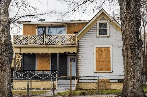 An old house with boarded up windows is surrounded by trees.