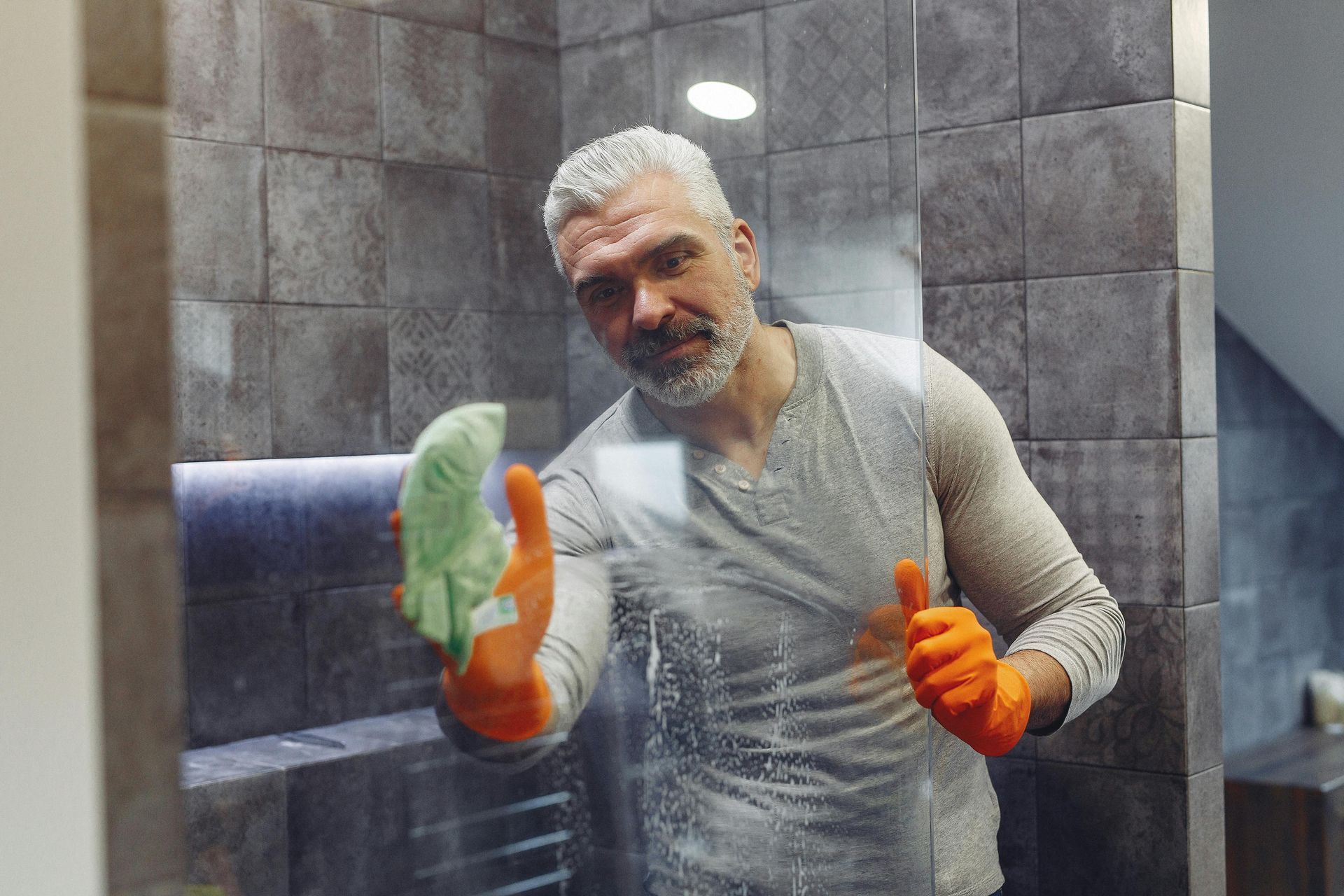A man is cleaning a bathroom mirror with a cloth and giving a thumbs up.