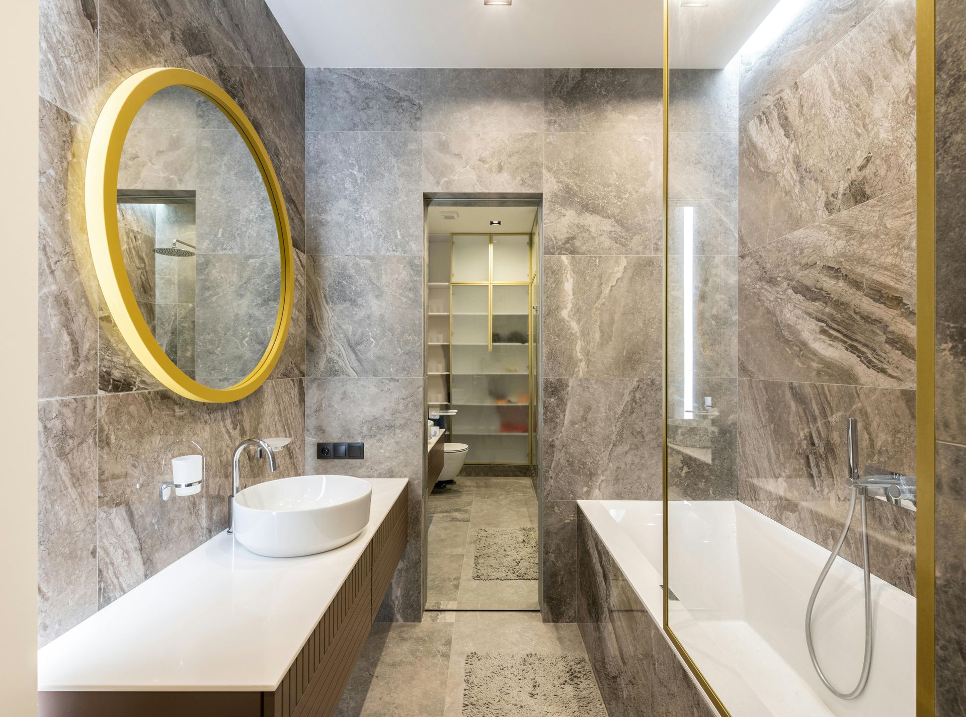 Modern bathroom with marble walls, round gold mirror, white sink and tub, and walk-in closet.